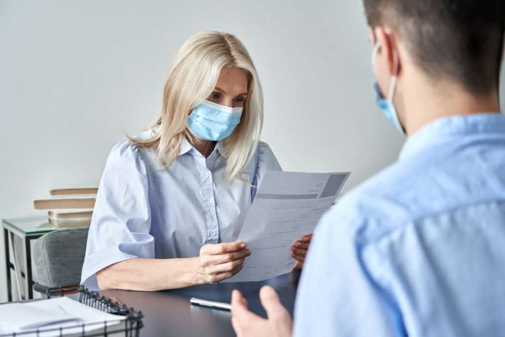 Female hr manager, employer wearing face mask reading cv at job interview.